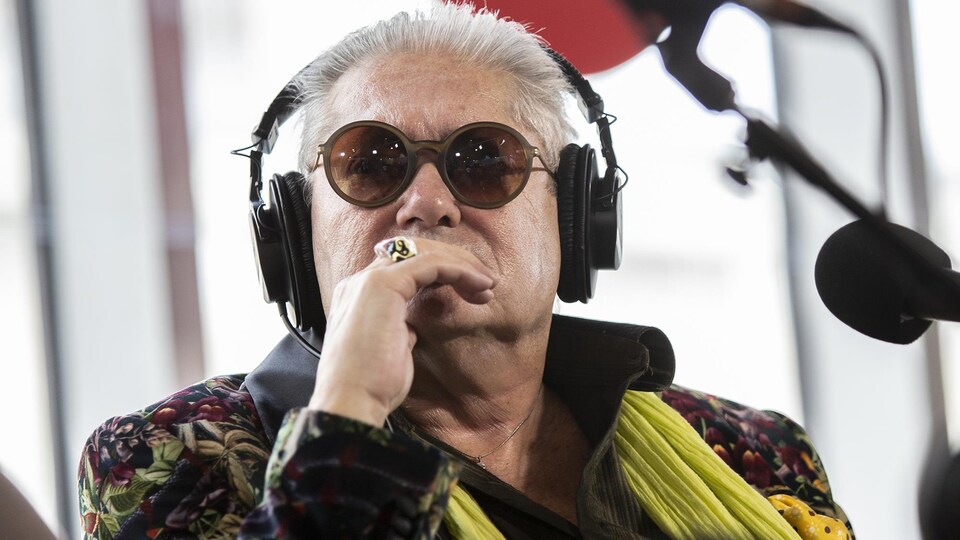 Un homme portant des écouteurs et des lunettes fumées porte sa main devant sa bouche.