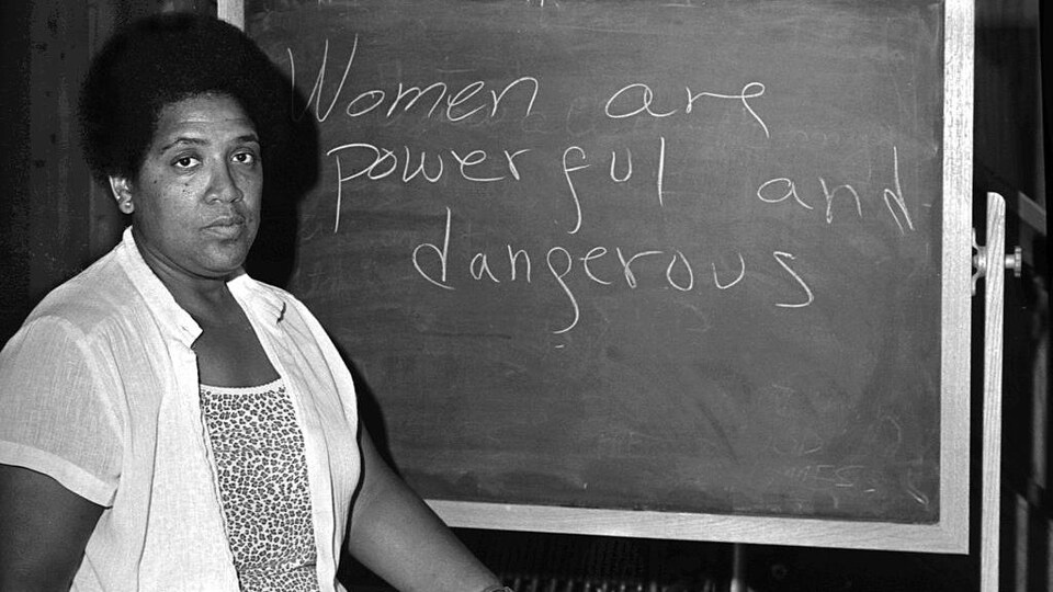 La poète noire pose devant un tableau où est inscrite la phrase : «Women are Powerful and Dangerous»