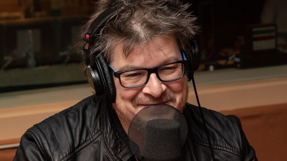 Un homme portant un casque d'écoute et des lunettes sourit devant un micro dans un studio de radio.