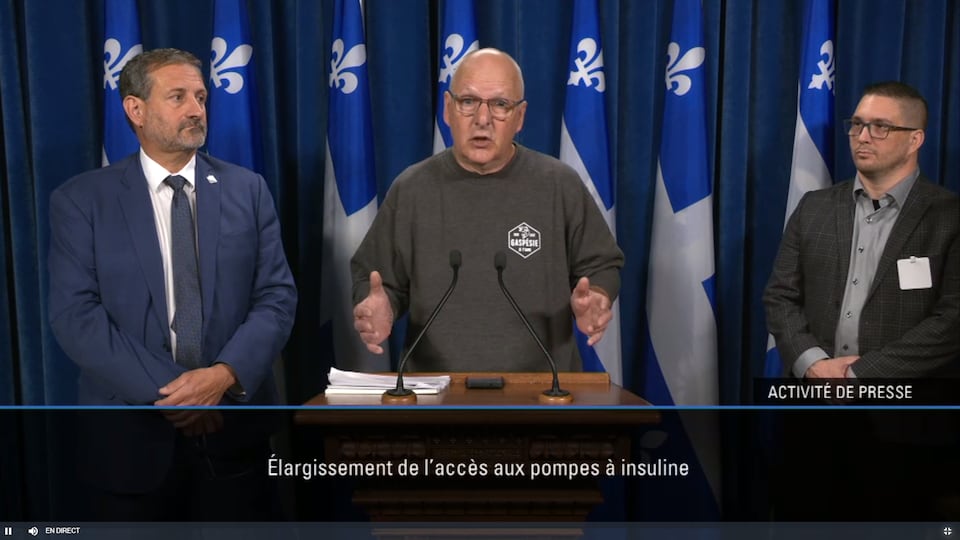 Les trois personnes côte-à-côte se tiennent devant plusieurs drapeaux du Québec en conférence de presse.