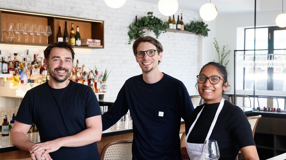 Deux hommes et une femme portant un tablier se tiennent debout dans une salle à manger de restaurant.