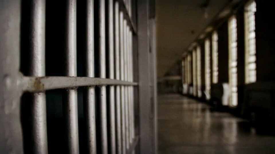 Le corridor d'une prison avec à gauche un gros plan sur les barreaux d'une cellule.