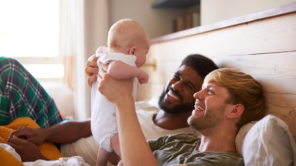 Deux hommes sont allongés dans un lit et sourient à un bébé de quelques mois.