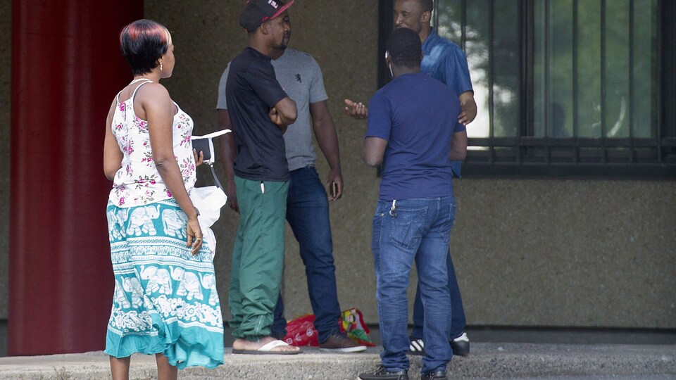 Quatre réfugiés parlent devant une maison. Une femme un peu à l'écart les regarde.