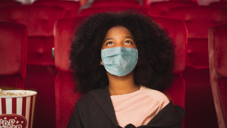 Une jeune fille portant un masque de protection est assise dans une salle de cinéma, avec un popcorn.