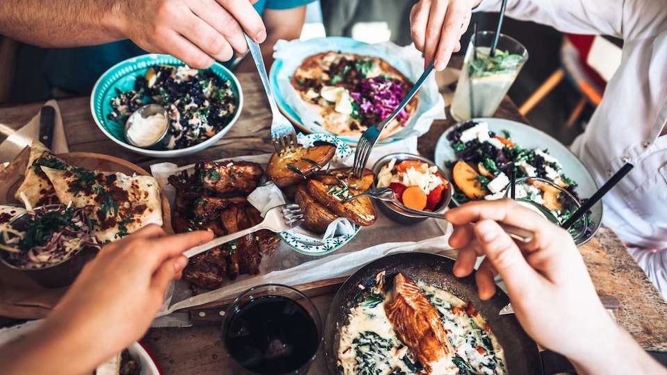 Plusieurs assiettes remplies de nourritures sont sur une table autour desquelles se trouvent des gens se servant de leurs fourchettes.