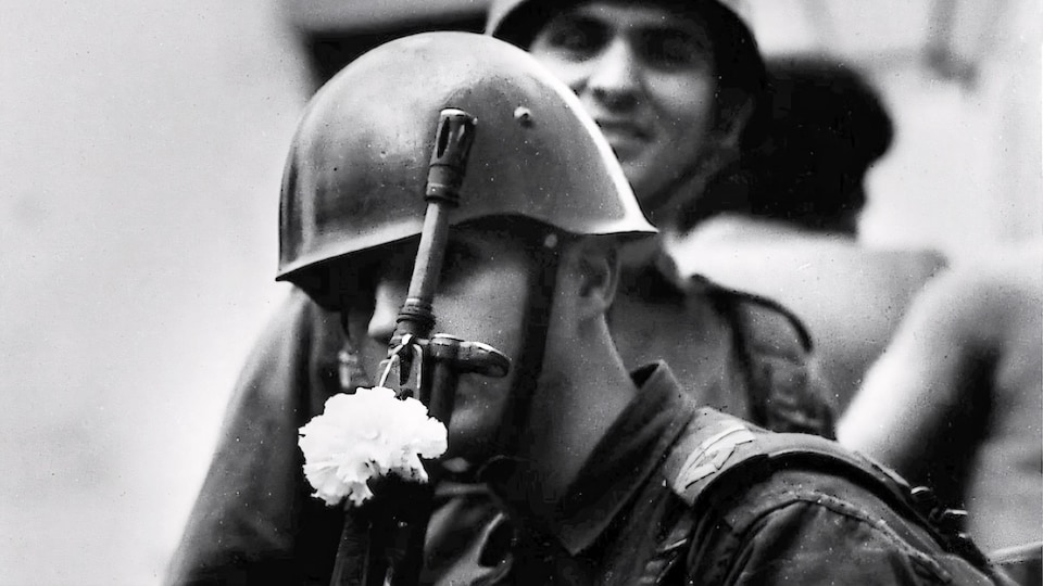 Plan rapproché en noir et blanc d'un soldat qui tient un fusil dans lequel est inséré une fleur blanche.