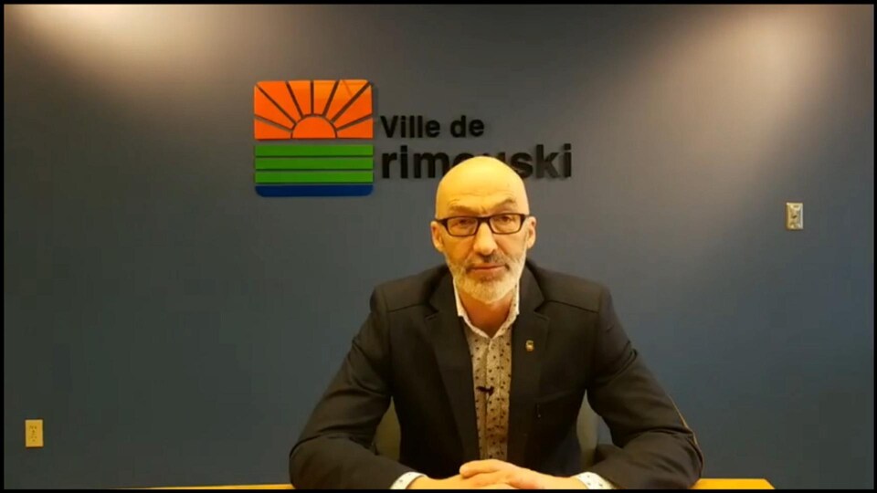 Un homme assis les mains croisées devant lui s'adresse à la caméra.
