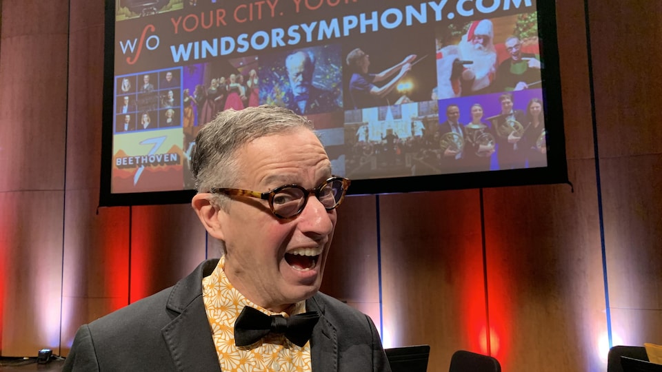 Robert Franz fait un grand sourire. Il porte des lunettes et il est debout devant un écran qui montre un montage de photos de l'Orchestre symphonique de Windsor.
