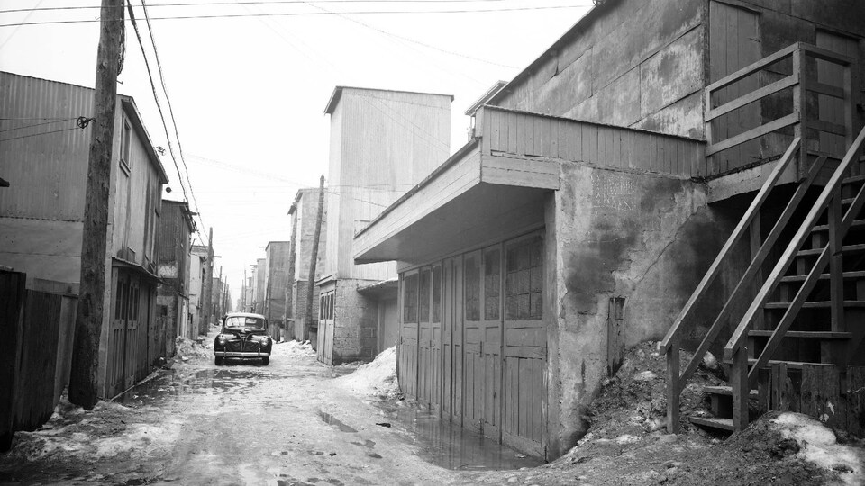 Photo en noir et blanc montrant une ruelle avec une voiture des années 40, bordée de bâtiments de part et d'autre.