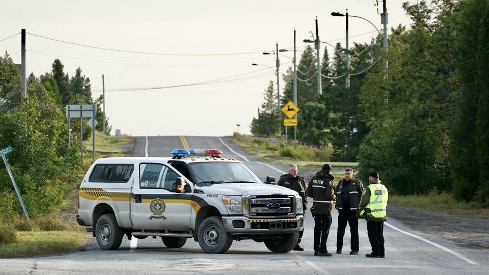 Une voiture de police stationnée au travers d'une route de campagne. Quatre policiers discutent dehors.