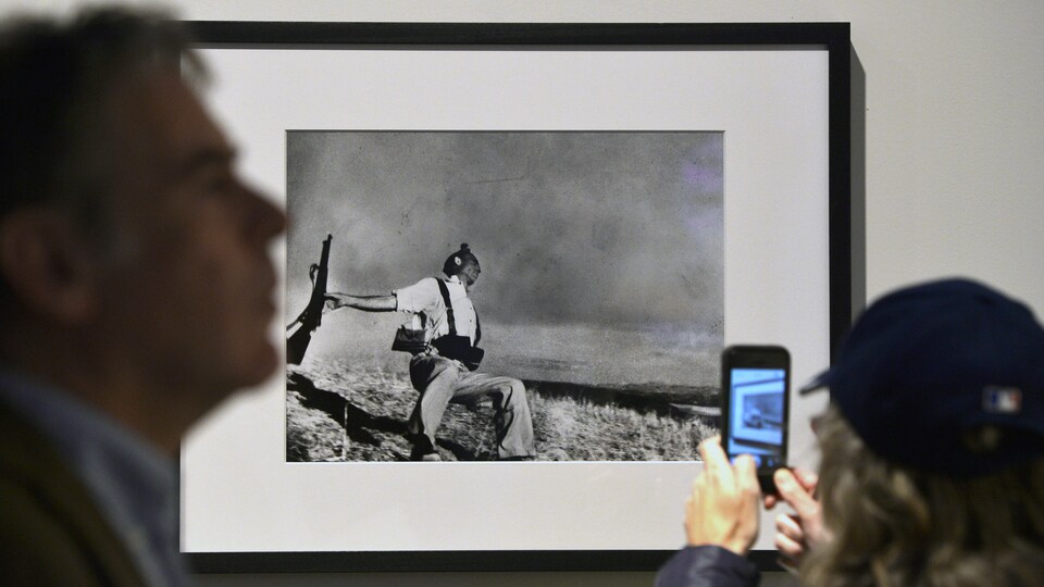 Une femme, de dos, tient un téléphone intelligent dans ses mains sur l'écran duquel on voit la photo encadrée qui est accrochée au mur. Cette photo représente un soldat semi accroupi et une carabine à la main.