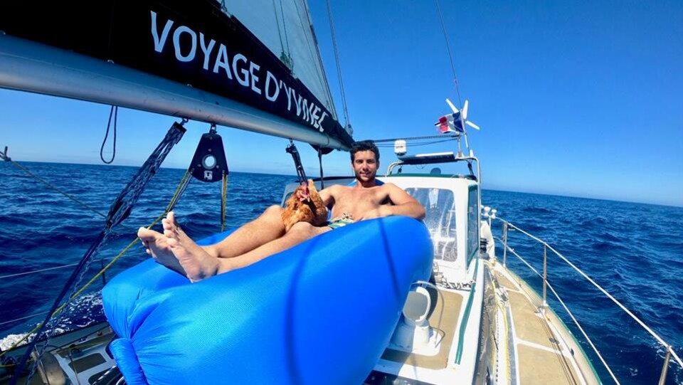 Le navigateur, torse nu, est confortablement installé sur une structure gonflable sur son bateau, caressant sa poule Monique