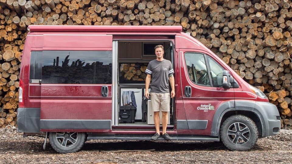 Le voyageur pose devant sa camionnette rouge, stationnée aux abords d'une immense corde de bois
