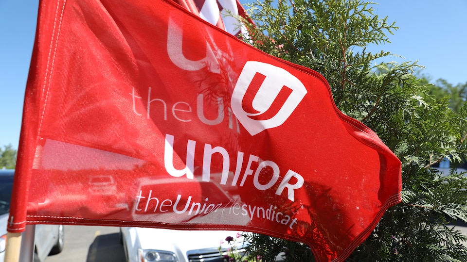 Un drapeau rouge, avec l'inscription Unifor en blanc, devant un tuya et des voitures.