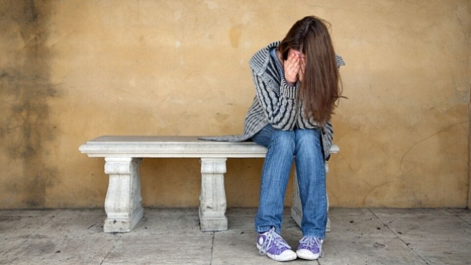 Une jeune fille se tient le visage entre les mains, elle est assise sur un banc.