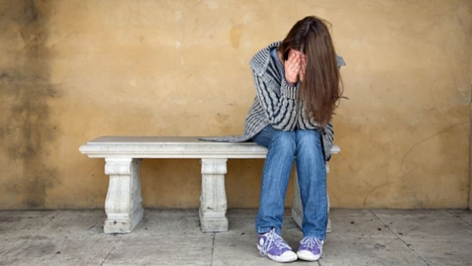 Une jeune fille se tient le visage entre les mains, elle est assise sur un banc.