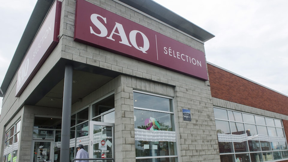 Un homme, en chemise et en pantalon, sort d'un bâtiment. Sur l'enseigne du bâtiment, on peut lire « SAQ Sélection ». 