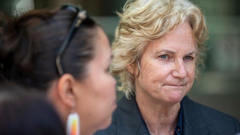 Une femme d'une quarantaine d'années aux cheveux courts et blonds. Elle regarde au loin avec un sentiment de perplexité.