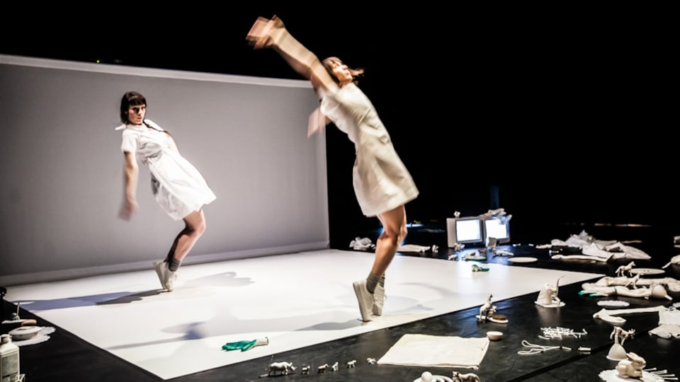 Deux interprètes en danse vêtues de blanc, sur une scène où se trouve plusieurs accessoires de couleur blanche.