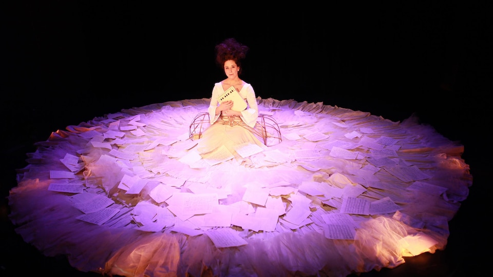 La comédienne Sofi Lambert, assise à genoux sur scène, tient un petit instrument de musique contre sa poitrine. Elle porte une immense robe sur laquelle il y a plusieurs feuilles de papier.