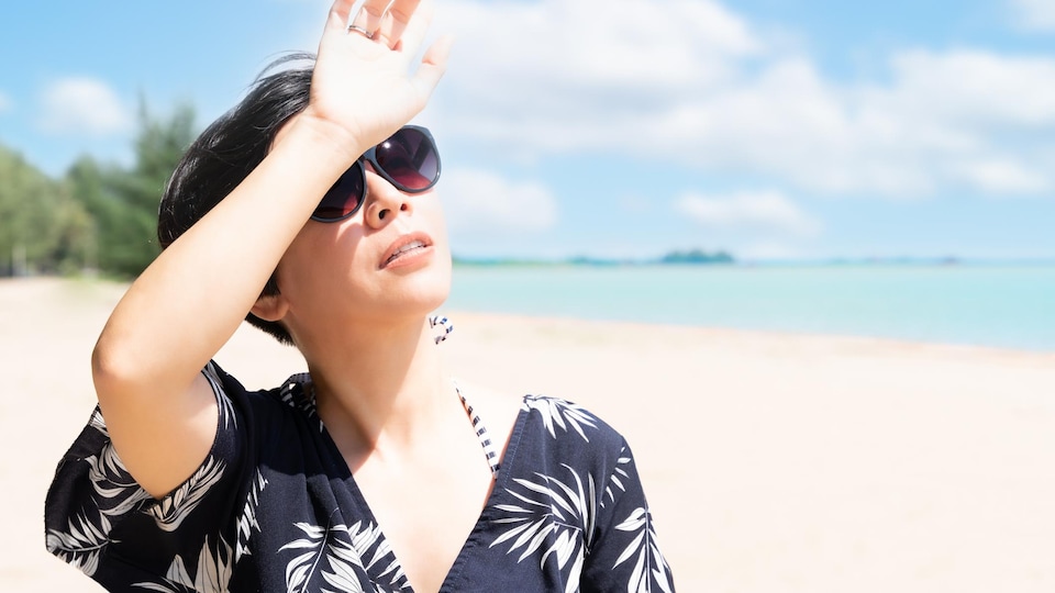 Une femme est sur la plage et elle regarde le soleil en se mettant la main devant les yeux.