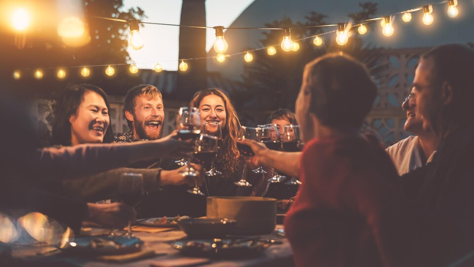 Des amis se font un toast avec des coupes de vin. Ils sont tous assis autour d'une table à l'extérieur, en début de soirée.