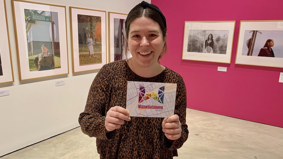 Sophie Hinch d'Art Windsor Essex est debout dans une galerie d'art et tient une carte postale sur laquelle est écrite 'Waawiiatanong Forever'. Des photos de 20 pouces par 30 pouces l'entourent.