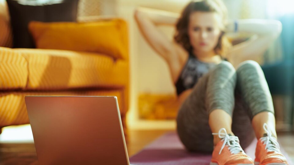 Une femme fait des abdos sur un tapis de yoga dans son salon en regardant un écran d'ordinateur.