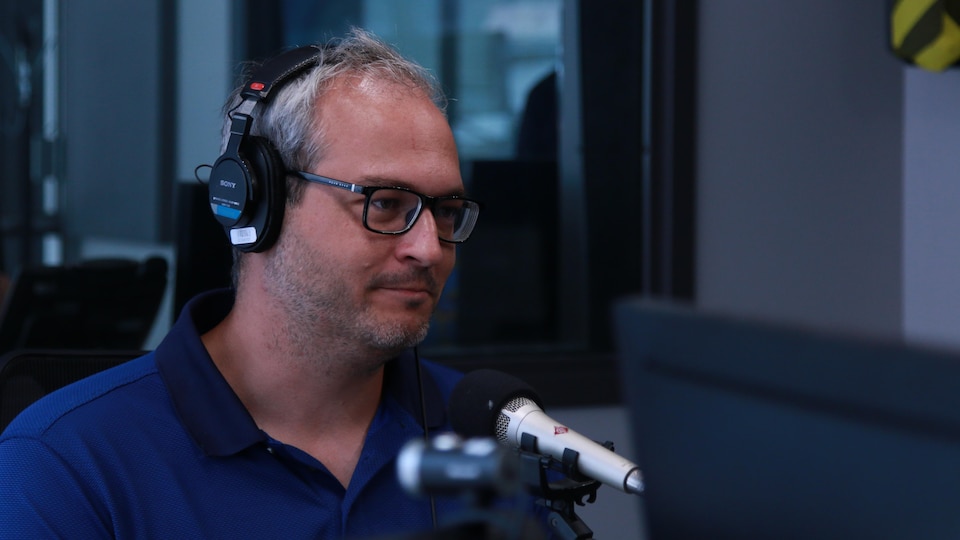 Une homme grisonnant avec des lunettes et un casque d'écoute dans un studio de radio.