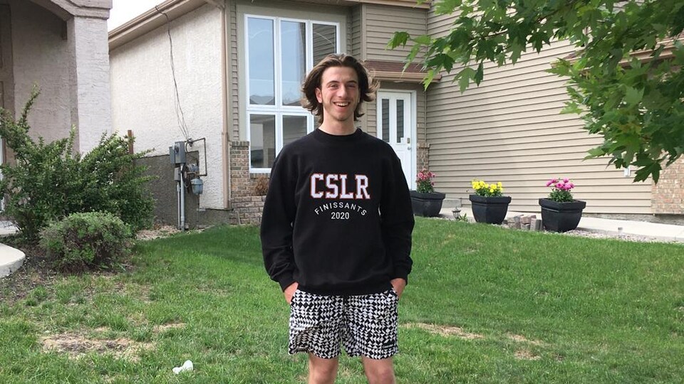 Un jeune homme au long cheveux et au visage souriant se tient dans le gazon devant une maison