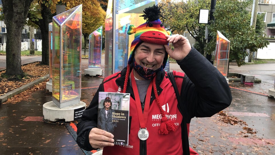 Stéphane Scott tient dans sa main son agenda Hope in Shadows 2021. Il a le manteau rouge des vendeurs Megaphone, une médaille au coup et un chapeau multicolore.