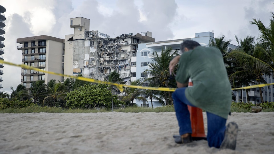 Un homme se recueille sur la plage, près du périmètre de sécurité. Devant lui, on aperçoit les restes de l'immeuble effondré.
