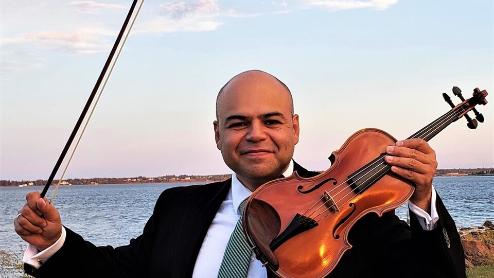 Un homme tient un violon et sourit à la caméra avec une rivière en arrière-plan.