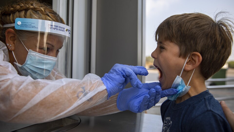Une infirmière avec une visière et des gants administre un test covid à un jeune garçon en introduisant un écouvillon dans sa bouche.