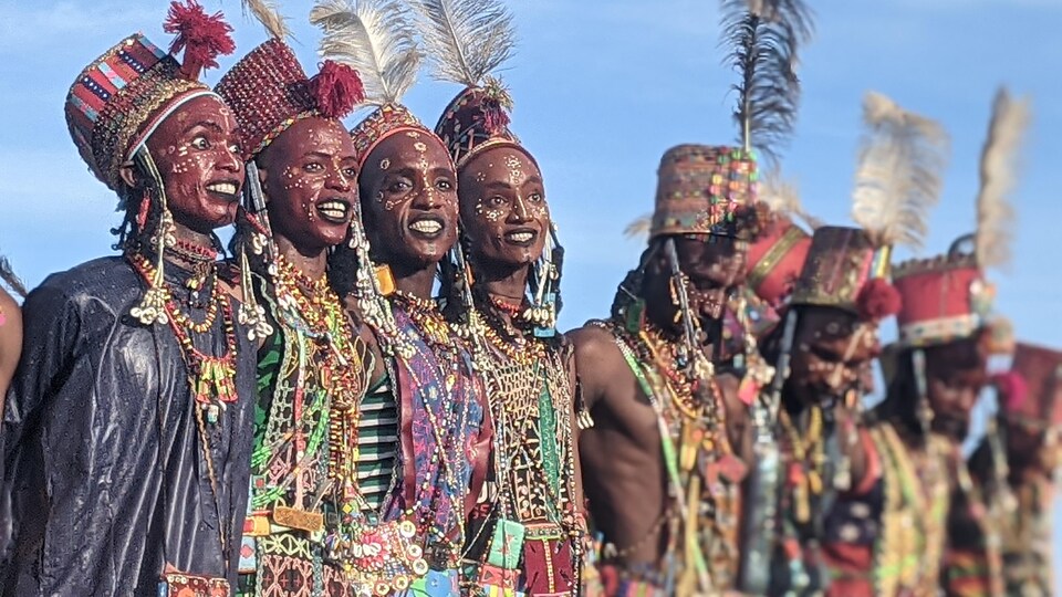 Quatre femmes souriantes en habits traditionnels du peuple Wodaabé en premier plan. À côté d'elles, des hommes du même peuple nomade en habits traditionnels.