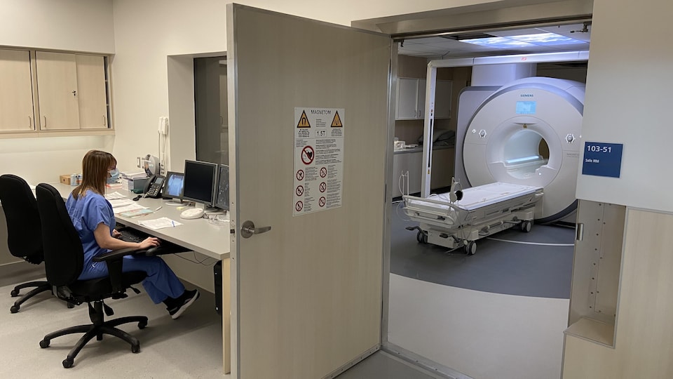 Une technologue en imagerie médicale travaille à l'ordinateur de l'autre côté d'une fenêtre qui donne accès à l'appareil d'IRM.