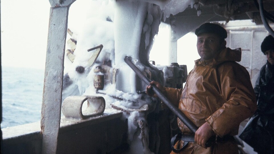 Un marin à l'imperméable jaune regarde l'objectif et tient une barre de métal dans ses mains nues. Autour de lui, la structure du navire est en glace.