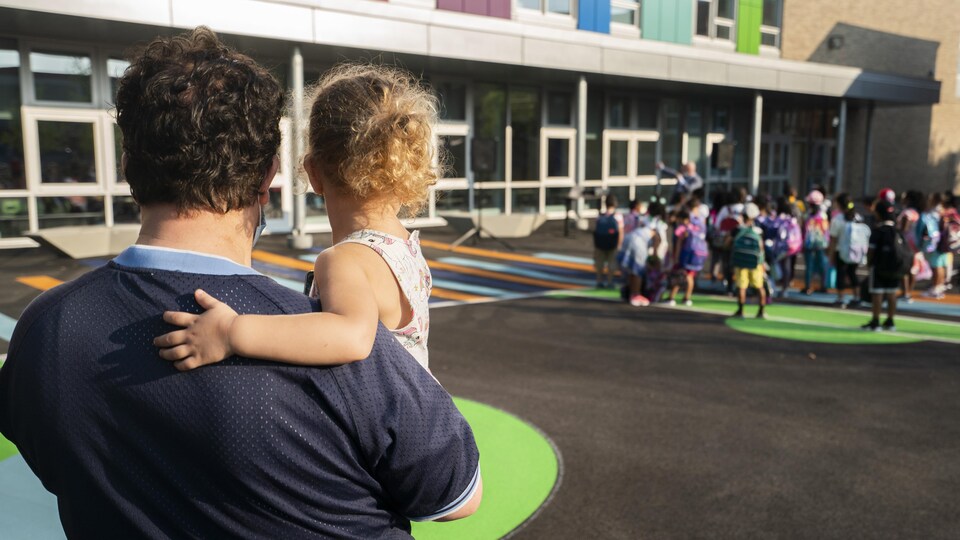 Un parent tient un enfant dans les bras et observe des élèves du primaire dans une cour d’école dans le quartier Saint-Michel.