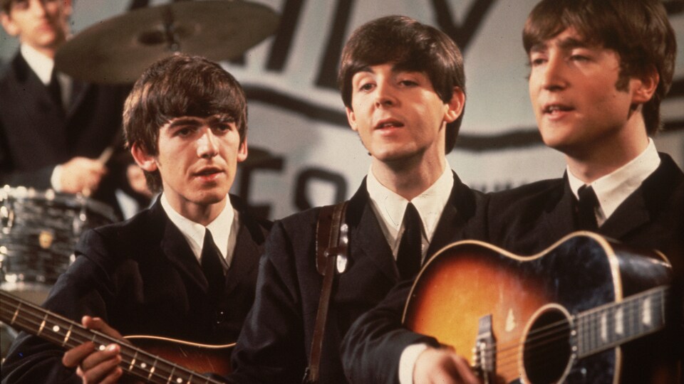 Une photo en couleurs des années 1960 montre les Beatles en complet noir pendant une performance.