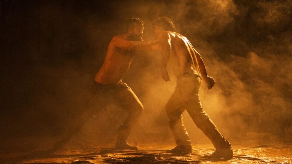 Deux hommes torse nu se bousculent sur une scène de théâtre enfumée.