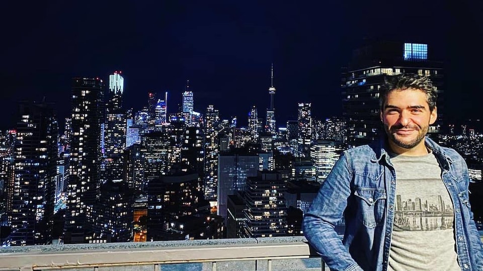 Un homme sourit sur un terasse du centre-ville de Toronto qui surpombe des gratte-ciels.