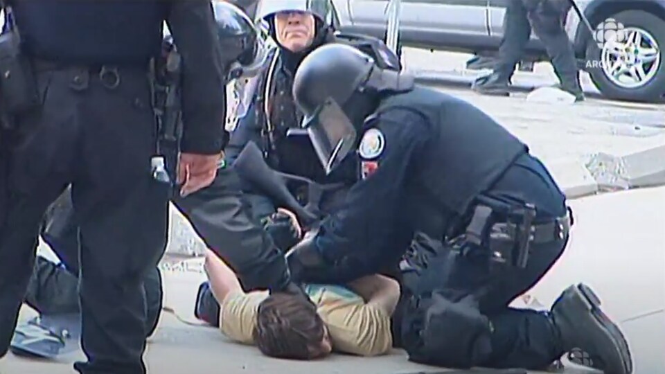 Un manifestant à bicyclette tombe par terre devant des policiers tenant des matraques et des boucliers de protection. 