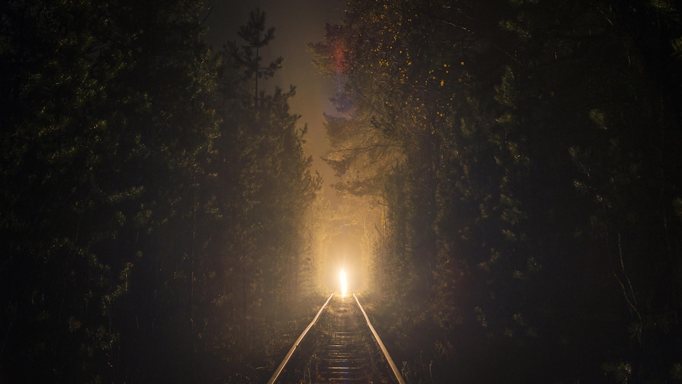 Une lumière au bout du chemin de fer qui traverse la forêt en pleine nuit.