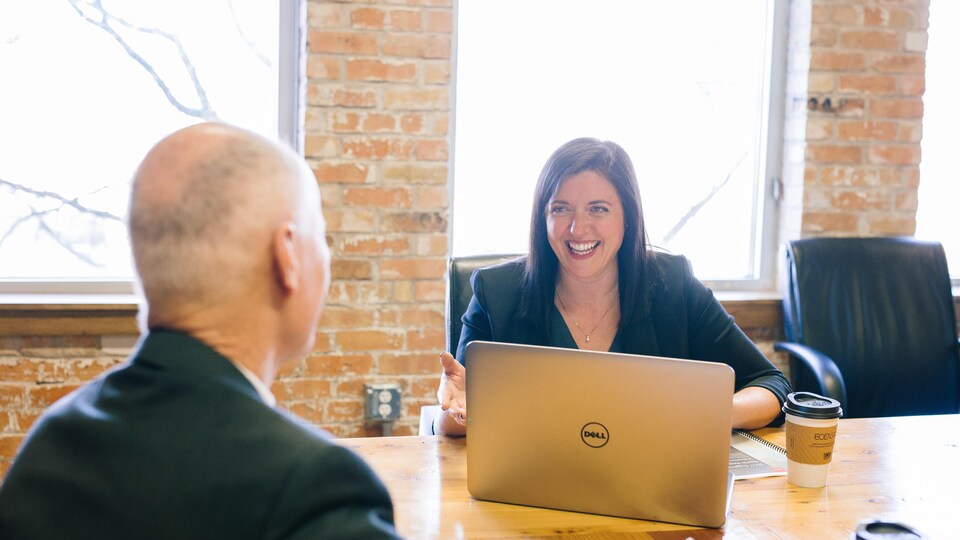 Une femme devant un ordinateur sourit à un collègue assis en face d'elle.