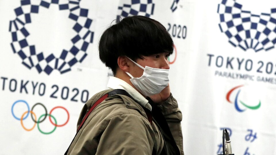 Un homme d'origine asiatique porte un masque sanitaire. Derrière lui, un bandeau fait la promotion des Jeux olympiques de Tokyo.