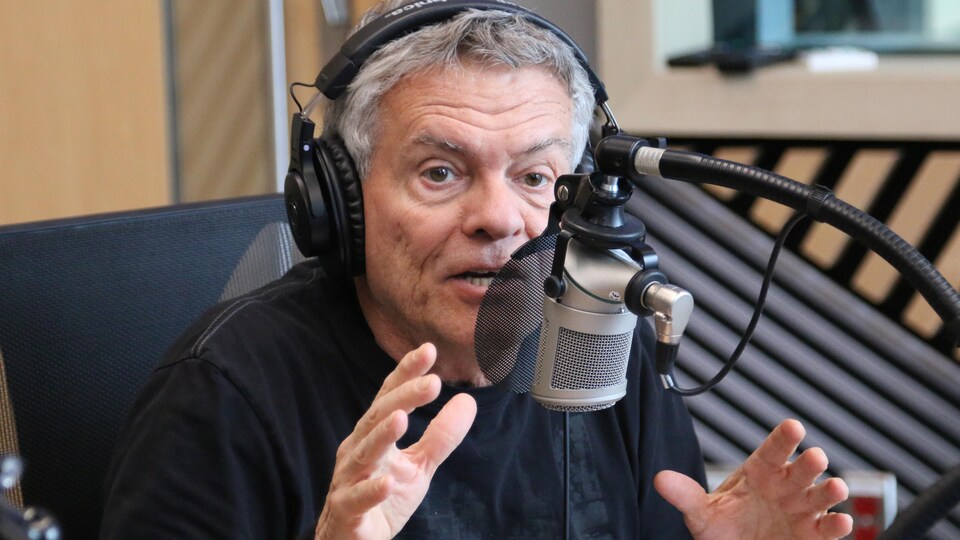 Un homme, casque d'écoute sur la tête, parle dans un micro dans un studio de radio tout en gesticulant. 