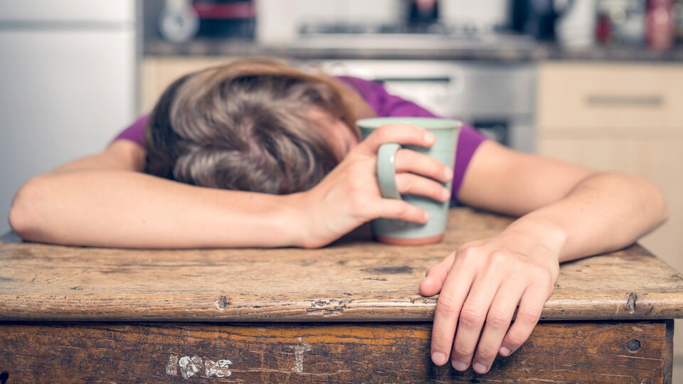 Une femme fatiguée se repose sur une table avec une tasse dans la main.