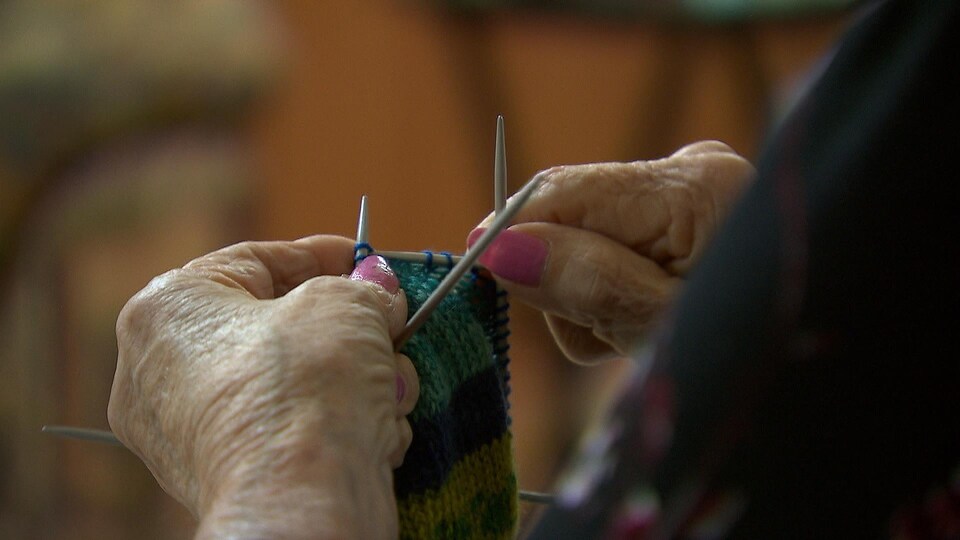 Plan rapproché sur des mains d'une personne âgée en train de tricoter. Quatre crochets de tricot permettent de tricoter la laine. 