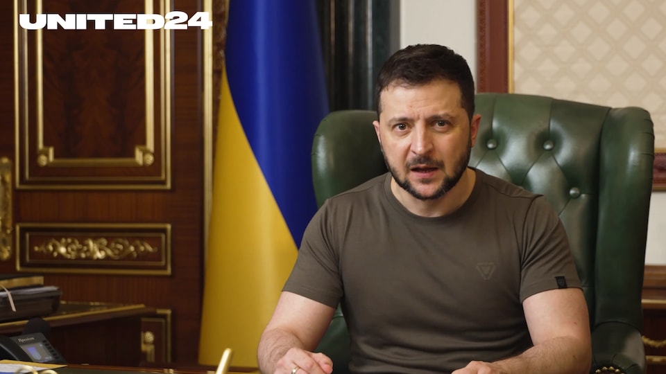 Volodymyr Zlensky s'adresse à la caméra, assis dans un bureau présidentiel, avec un drapeau de l'Ukraine en arrière-plan. 
